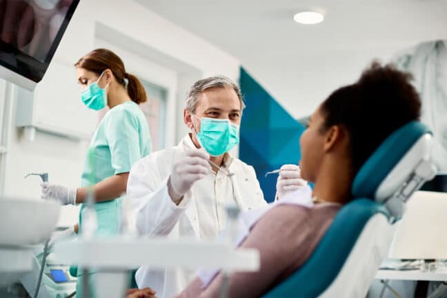 A dentist about to start an exam on a patient seated in a dental chair, with a dental assistant standing in the background.