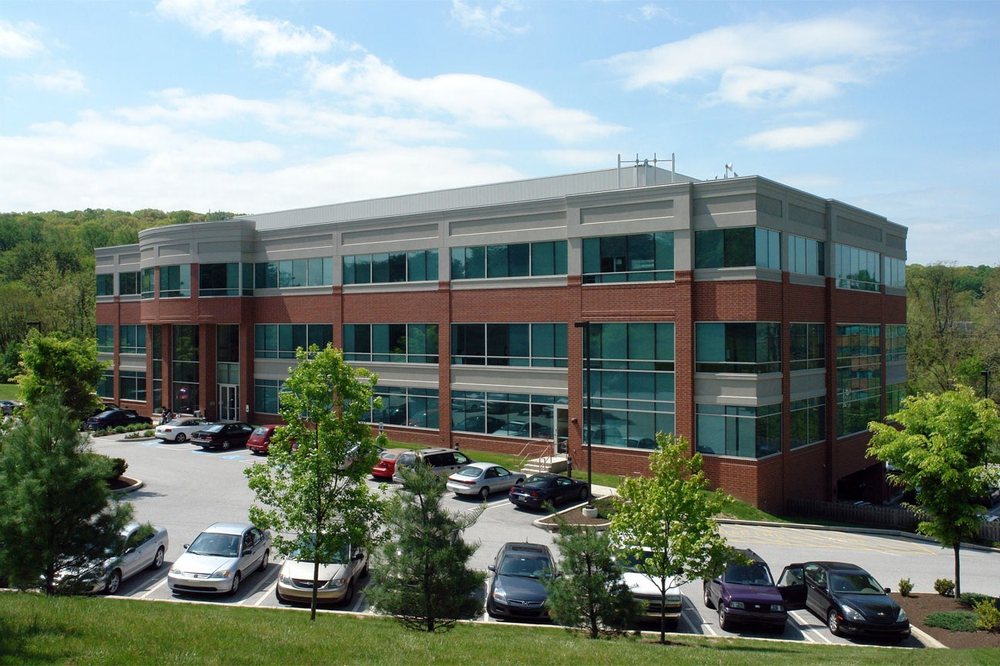 Modern three-story brick and glass office building with large windows, surrounded by trees and a parking lot, representing a dental organization’s administrative or clinical headquarters.